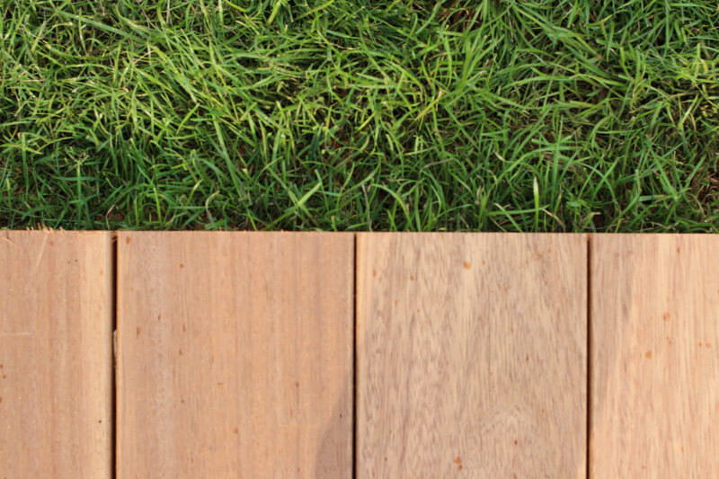 Eine edle Holzterrasse grenzt direkt an den saftig grünen Gartenrasen und einen schmalen Gartenweg an.